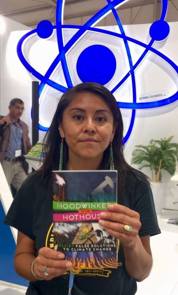 Leona Morgan of NM No False Solutions Coalition, holding "Hoodwinked in the Hothouse" book in front of nuclear symbol