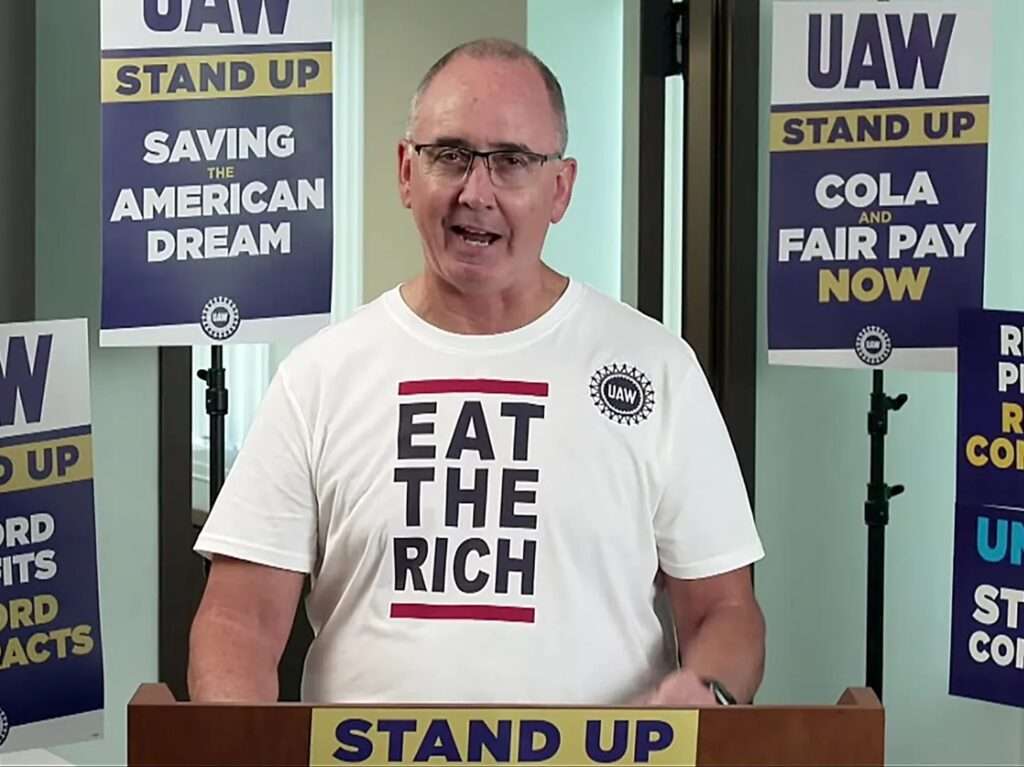UAW President Shawn Fain at podium with picket signs and t shirt proclaiming "Eat the Rich"